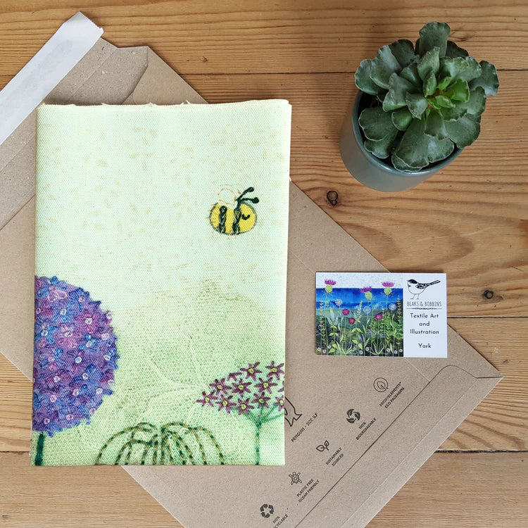 Embroidered fabric with a bee and flower design on a wooden surface with a plant and card.