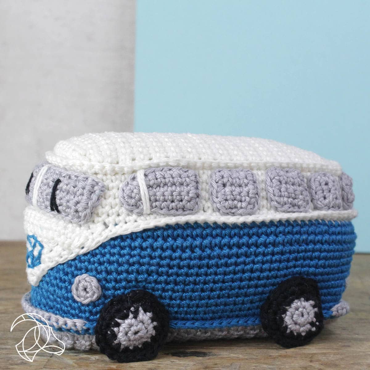 Crocheted blue and white van toy on a wooden surface with a light blue wall in the background.