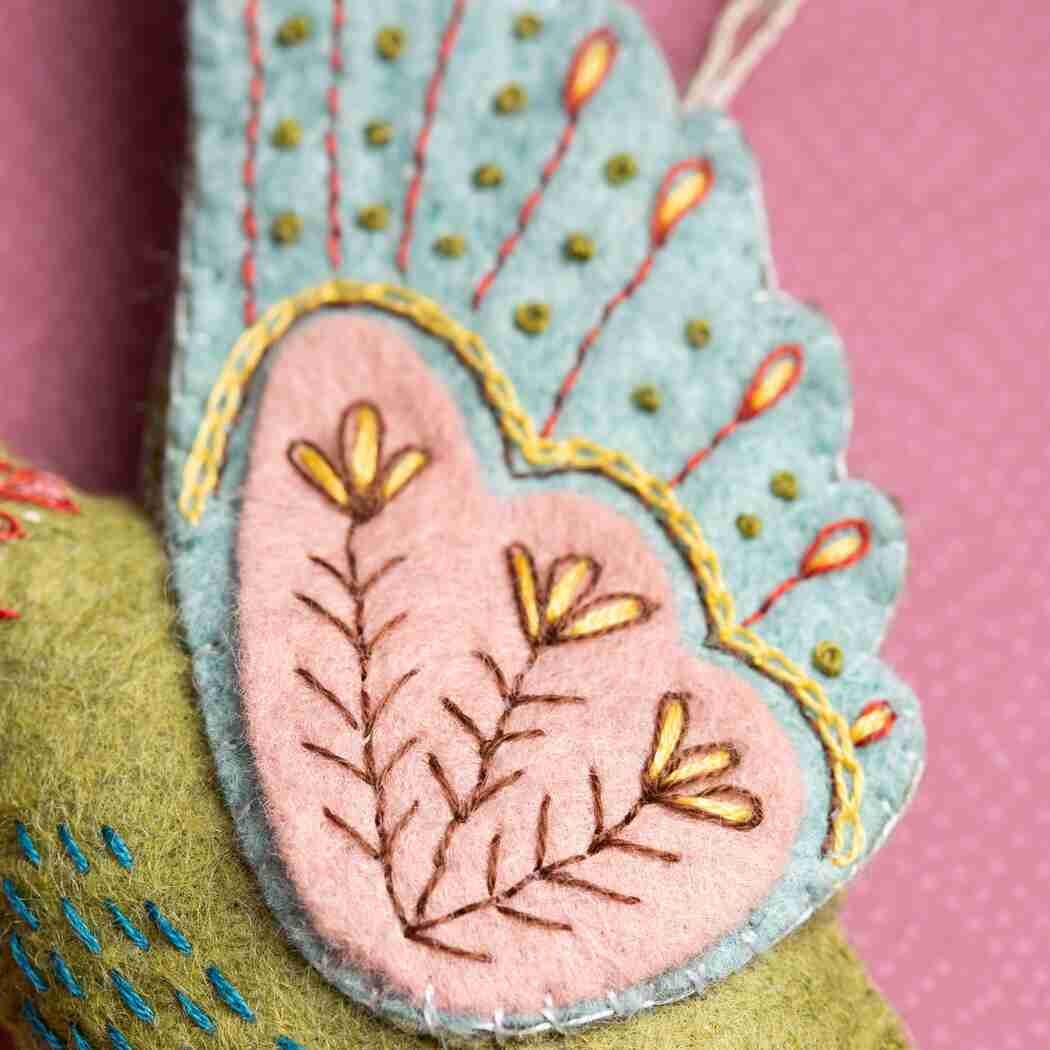 Close-up of a handmade felt bird wing ornament in light blue and pink with yellow and brown embroidered details, attached to a green felt bird body against a pink background.